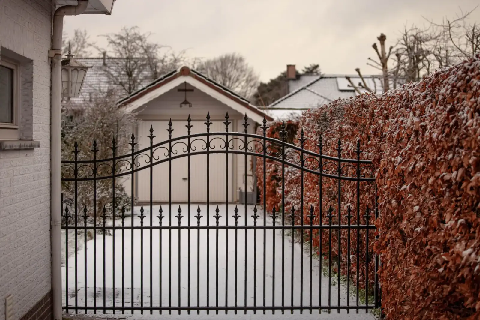 Black ornamental steel driveway gate with an arched top rail and spear-tipped pickets, closed across a snow-covered drive